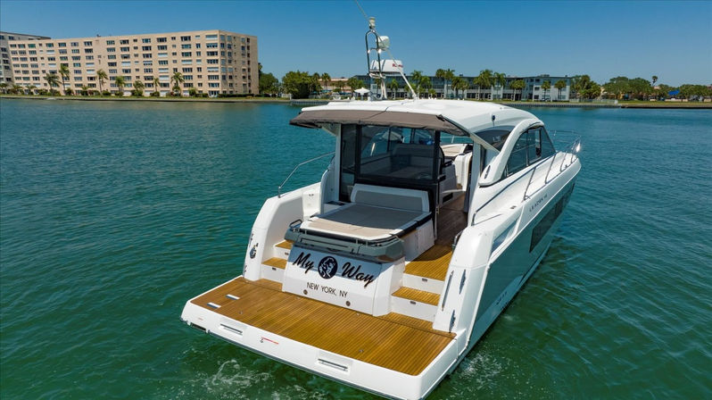 Sleek white motor yacht with teak swim platform cruising on clear turquoise water near palm-tree lined waterfront condos under a sunny blue sky.