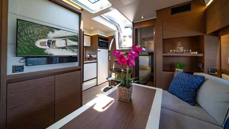 Interior of a modern motor yacht salon with warm wood cabinetry, compact galley and skylight, wall TV showing an aerial boat, dining table with bright pink orchids and cushioned seating with a blue throw pillow.