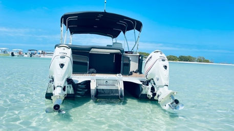 Rear view of a pontoon boat with twin outboard engines and a black bimini top anchored in crystal-clear shallow turquoise water near a sandy island under a bright blue sky.