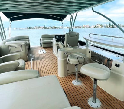 Luxury pontoon boat interior with teak-style deck, plush swivel bar stools, lounge seating and shaded canopy overlooking a calm bay and distant coastal skyline.