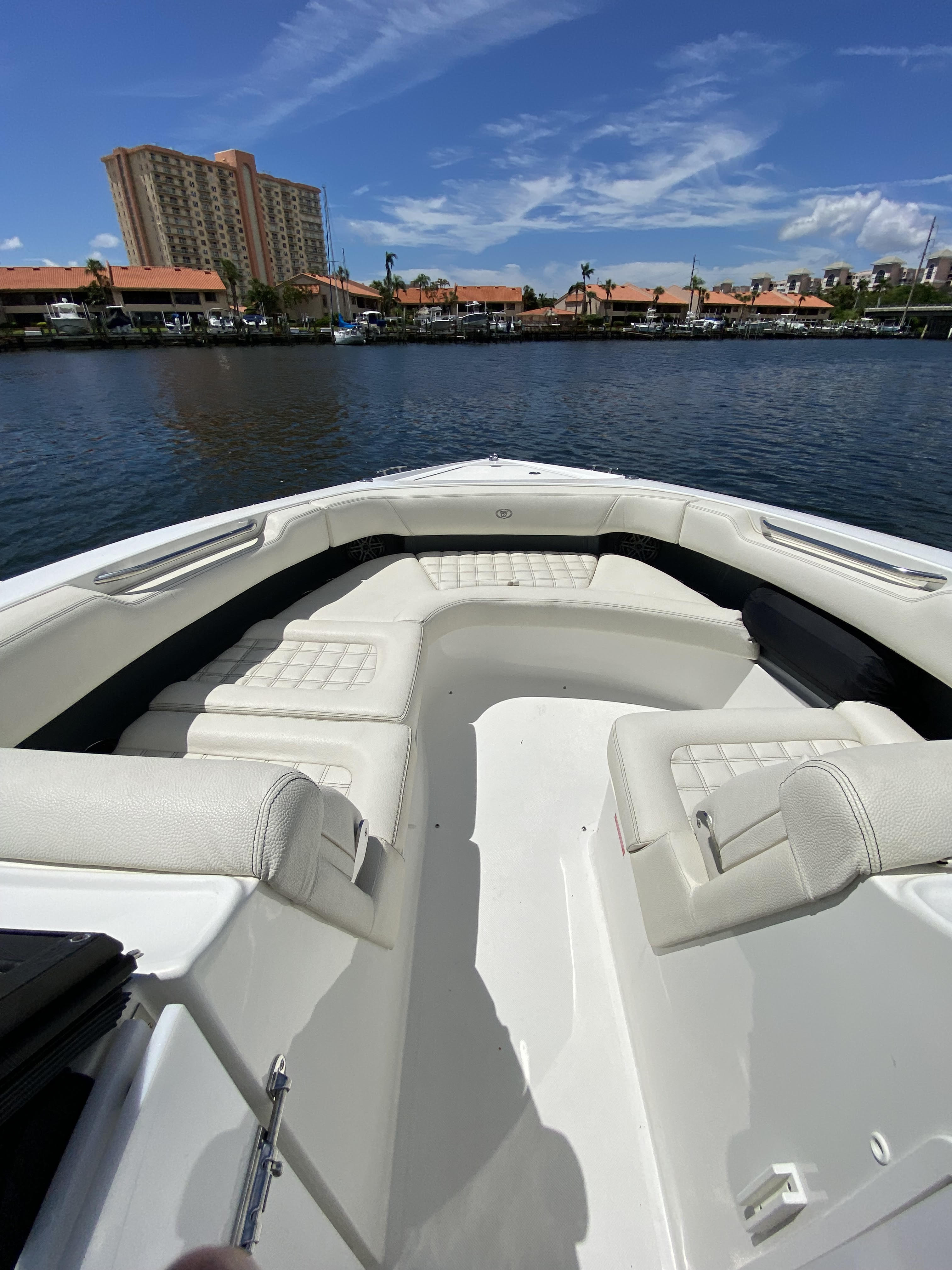 White powerboat bow with cushioned U-shaped seating looking over a calm marina with docks, moored boats and orange-roof waterfront condos under a bright blue sky.