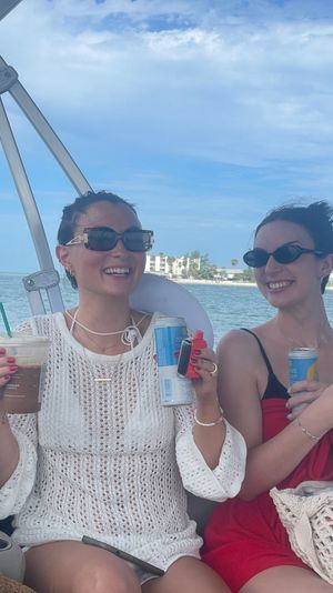 Two smiling friends in sunglasses enjoying drinks on a boat cruise — one in a white knit cover-up holding an iced coffee and a can, the other in a red dress, with turquoise ocean and beachfront buildings under a sunny blue sky.