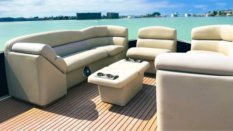 Beige leather lounge seating and center ottoman with cupholders on a teak-deck boat, turquoise coastal water and distant shoreline under a sunny blue sky.