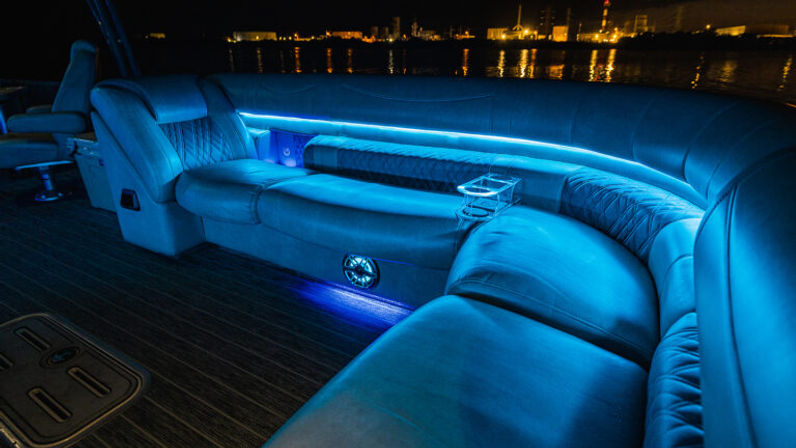Sleek curved blue-lit leather boat interior at night with LED underseat lighting, built-in cup holder and speaker, and waterfront city lights reflecting on calm water.