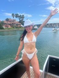 Smiling woman in white bikini and white cowboy hat reading 'BRIDE' posing on a boat in a sunny waterfront canal with palm trees, yachts and luxury homes