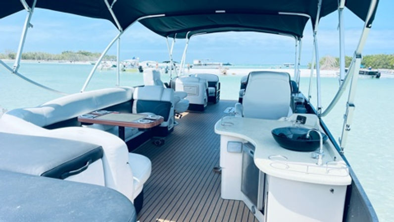 Pontoon boat interior with cushioned lounges, small table and wet bar under a canopy, anchored in shallow turquoise water by a sandy sandbar.
