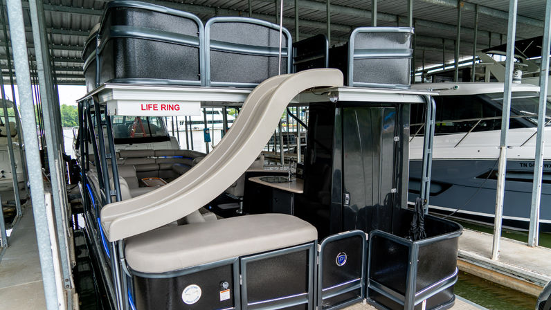 Party-ready pontoon boat with beige water slide, cushioned seating and small cabin, docked in a covered lake marina slip beside other yachts.