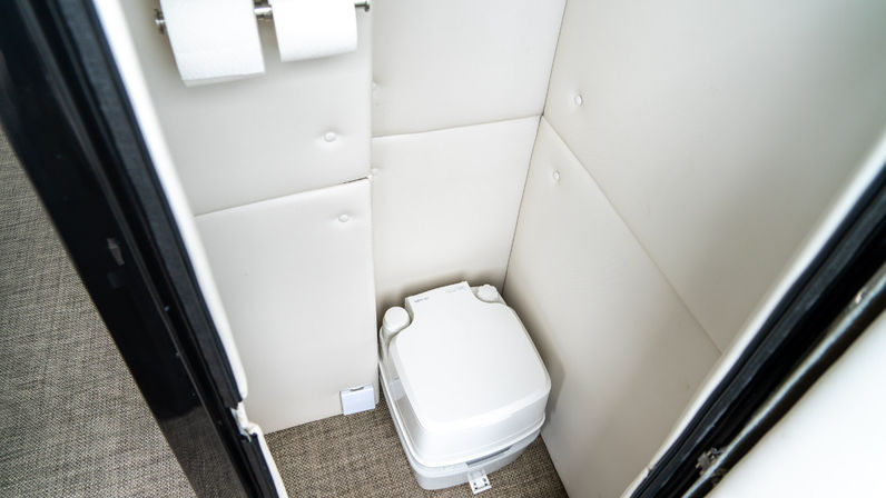 Compact white portable camper toilet in a small padded beige RV/boat bathroom with carpeted floor and two toilet paper rolls