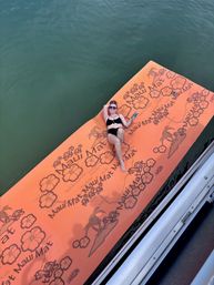 Person lounging on a large orange tropical-patterned floating mat with hibiscus prints, holding a drink while drifting on calm green water beside a boat.