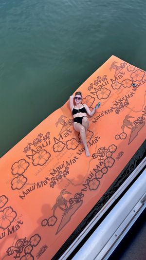 Person lounging on a large orange tropical-patterned floating mat with hibiscus prints, holding a drink while drifting on calm green water beside a boat.