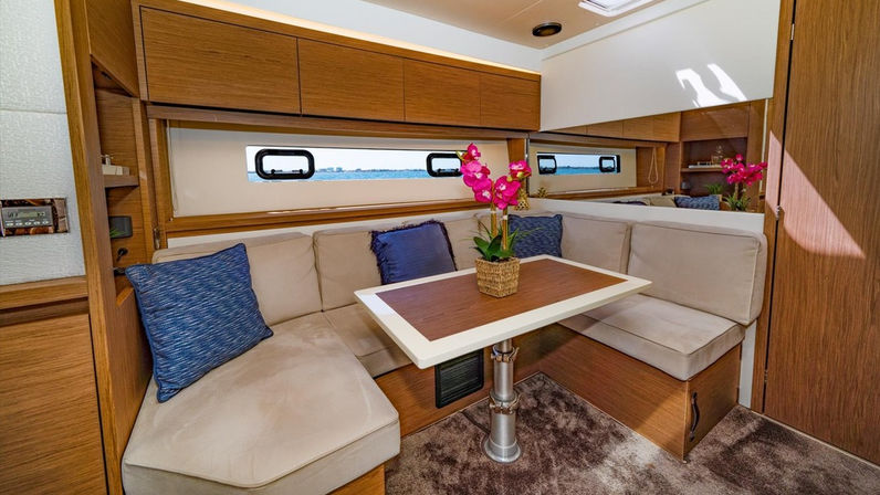 Cozy yacht salon dining area with L-shaped beige sofa, blue throw pillows, wood table topped by a pink orchid centerpiece, narrow porthole windows and warm wood cabinetry.