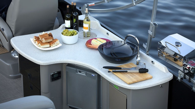 Pontoon boat galley kitchenette by calm water: curved countertop with sink and faucet, cutting board and tongs, bowl of grapes, bread and cheese plates, wine bottles and mini fridge—ready for a lakeside picnic.