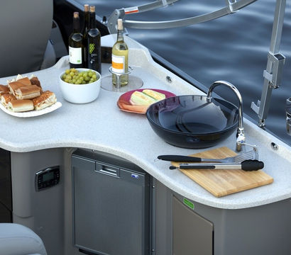 Pontoon boat galley kitchenette by calm water: curved countertop with sink and faucet, cutting board and tongs, bowl of grapes, bread and cheese plates, wine bottles and mini fridge—ready for a lakeside picnic.