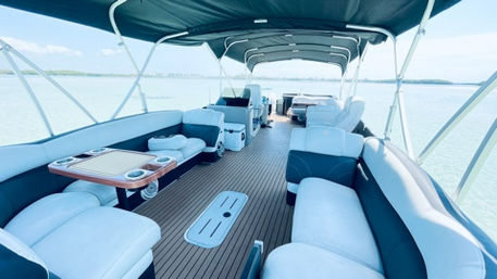 Spacious pontoon boat interior with wraparound white-and-teal cushioned seating, teak-style deck, center table with cupholders, overhead canopy and open view of calm blue water and distant shoreline — perfect for a relaxing lake or coastal cruise.
