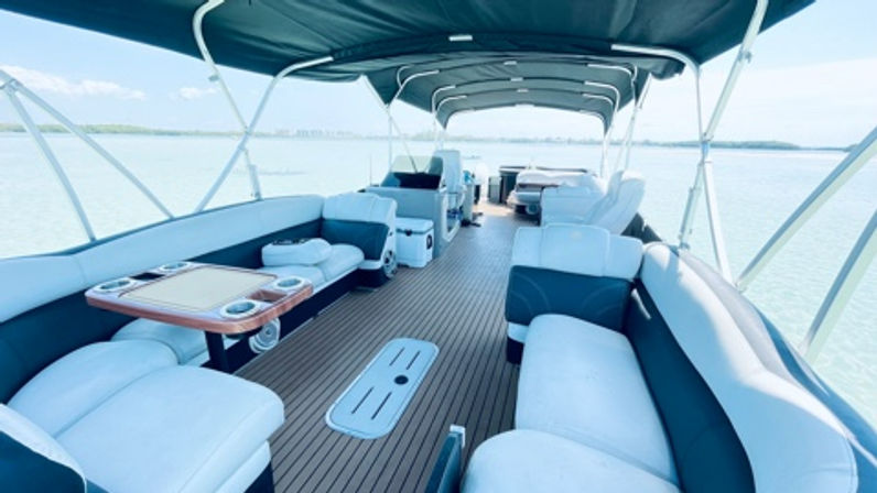 Spacious pontoon boat interior with wraparound white-and-teal cushioned seating, teak-style deck, center table with cupholders, overhead canopy and open view of calm blue water and distant shoreline — perfect for a relaxing lake or coastal cruise.