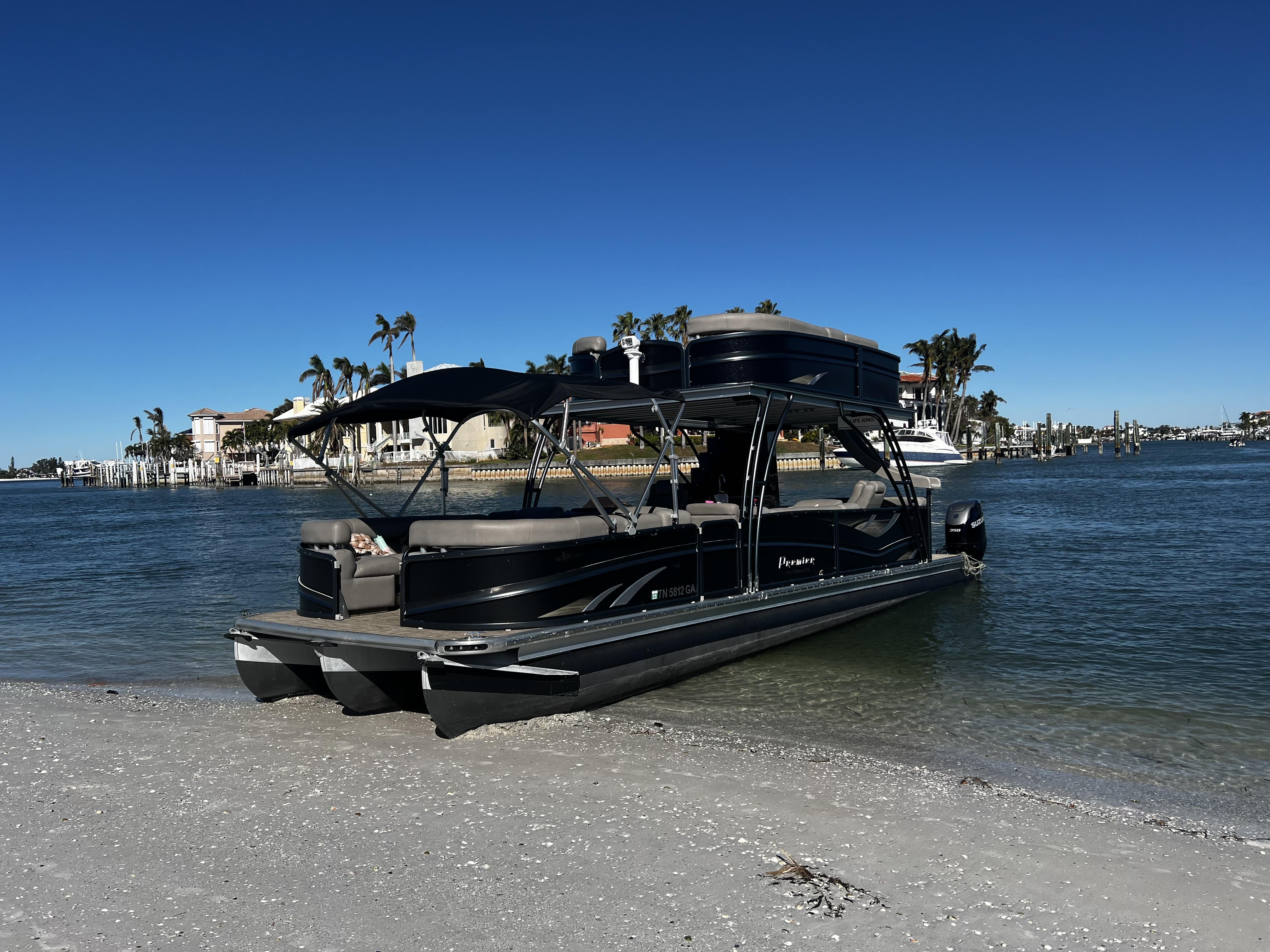 Luxury black pontoon boat beached on a sunny coastal shoreline with palm trees, waterfront homes, docks and a clear blue sky.