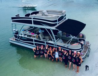Double-decker pontoon boat anchored in shallow clear water by a sandy shore with a group of women in matching black tank tops and pink sunglasses standing waist-deep — lively beach boat gathering.