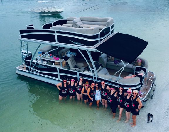Double-decker pontoon boat anchored in shallow clear water by a sandy shore with a group of women in matching black tank tops and pink sunglasses standing waist-deep — lively beach boat gathering.