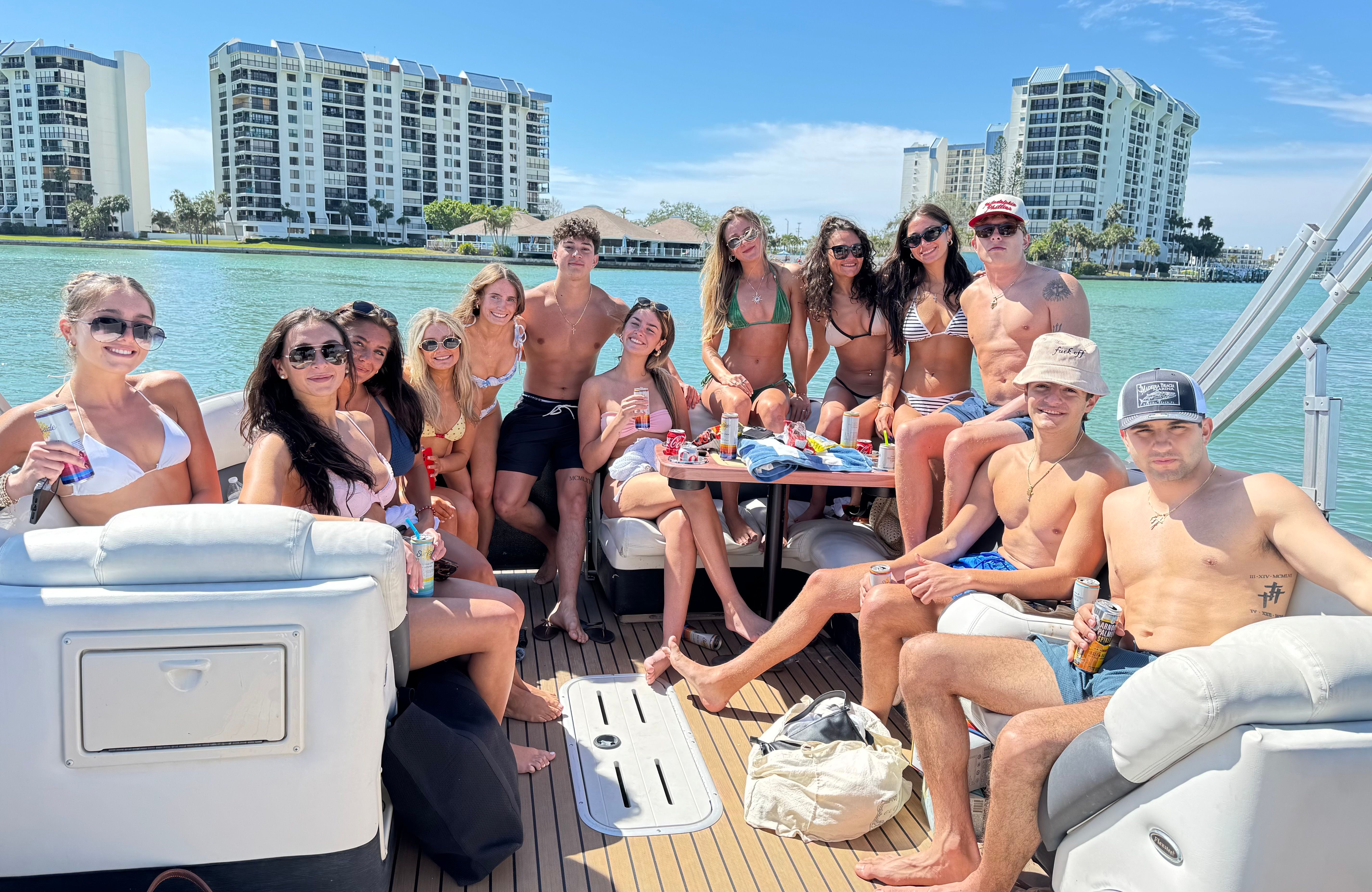 Smiling group of young adults in bikinis and swim trunks enjoying drinks on a pontoon boat, turquoise bay and waterfront condos on a sunny day