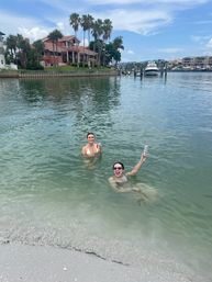 Two friends swimming in shallow bay water near a sandy shore, holding canned drinks and smiling, with palm trees, waterfront homes and a docked yacht under a blue sky