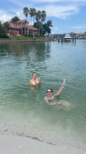 Two friends swimming in shallow bay water near a sandy shore, holding canned drinks and smiling, with palm trees, waterfront homes and a docked yacht under a blue sky