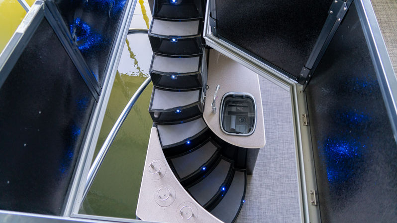 Sleek LED-lit curved boat stairs descending through an open hatch past a compact countertop and sink, with a glimpse of water below