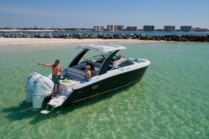 Sunlit motorboat with three people anchored in shallow turquoise water off a sandy beach and rock jetty, man pointing toward shore and low-rise coastal buildings on the horizon