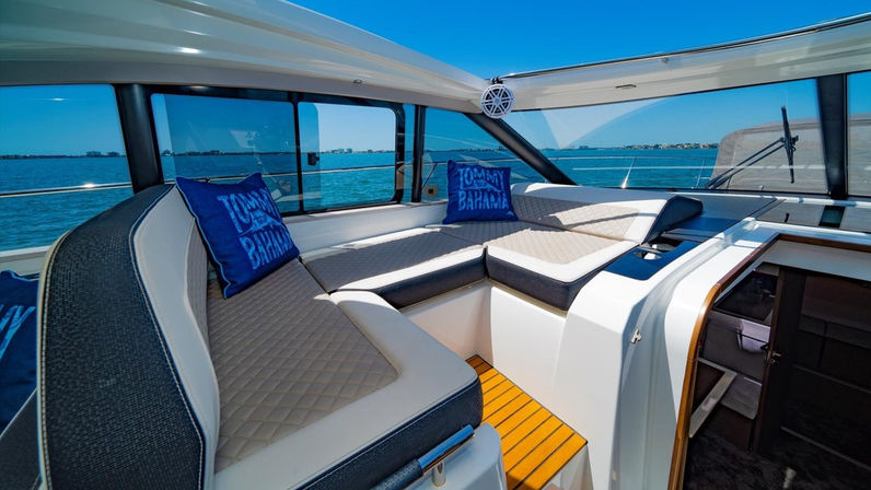 Sunlit yacht cabin with L-shaped quilted cream-and-gray seating, blue throw pillows, teak floorboard steps and panoramic windows framing turquoise coastal waters and a clear blue sky.