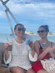 Two smiling friends in sunglasses enjoying drinks on a boat cruise — one in a white knit cover-up holding an iced coffee and a can, the other in a red dress, with turquoise ocean and beachfront buildings under a sunny blue sky.