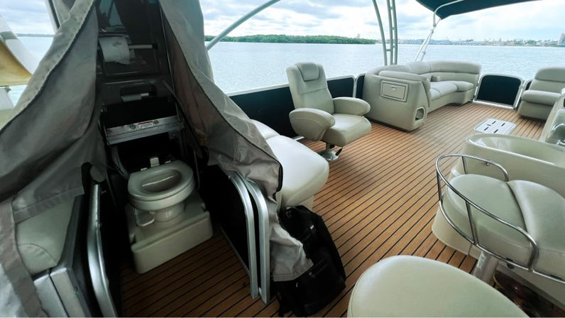 Pontoon boat deck with teak-style flooring, cream leather lounge seating and swivel chairs, small enclosed marine toilet visible behind a curtain, overlooking calm lake waters and distant shoreline.