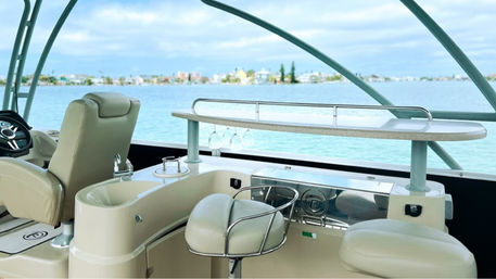Bay-view luxury boat helm and bar area with cream leather captain’s chair, swivel bar stools, small wet bar with hanging wine glasses, and a countertop overlooking turquoise coastal waters and a distant low-rise shoreline under a cloudy sky.