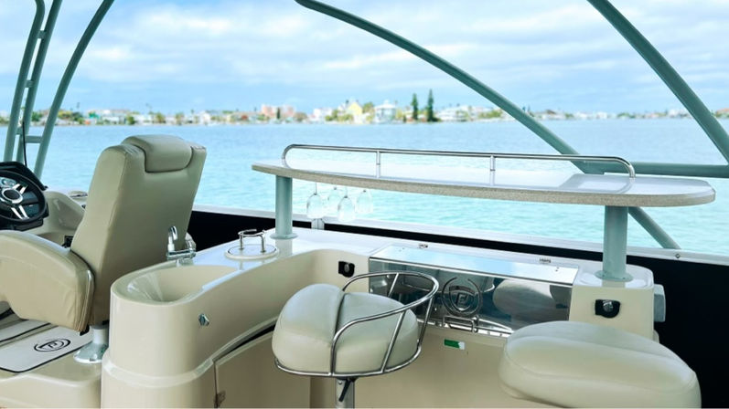 Bay-view luxury boat helm and bar area with cream leather captain’s chair, swivel bar stools, small wet bar with hanging wine glasses, and a countertop overlooking turquoise coastal waters and a distant low-rise shoreline under a cloudy sky.