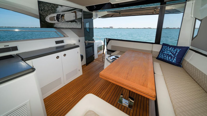 Sunlit motor yacht salon with teak flooring, wooden dining table, beige quilted bench seating and blue throw pillow, large windows revealing turquoise coastal waters.