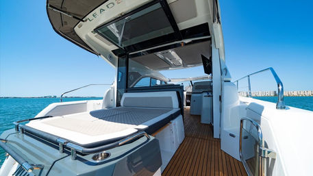 Sunlit rear deck of a luxury motor yacht with teak flooring, cushioned sunbed and stainless railings on clear turquoise coastal waters under a bright blue sky.