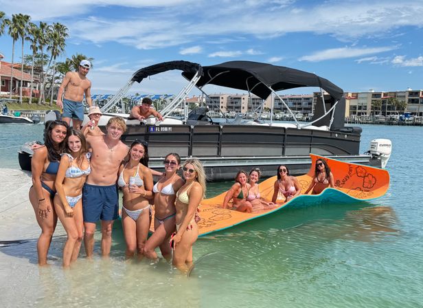 Smiling group of friends in colorful swimsuits enjoying shallow turquoise water beside a black pontoon boat and orange floating lounge at a sunny Florida beach marina with palm trees and waterfront condos.