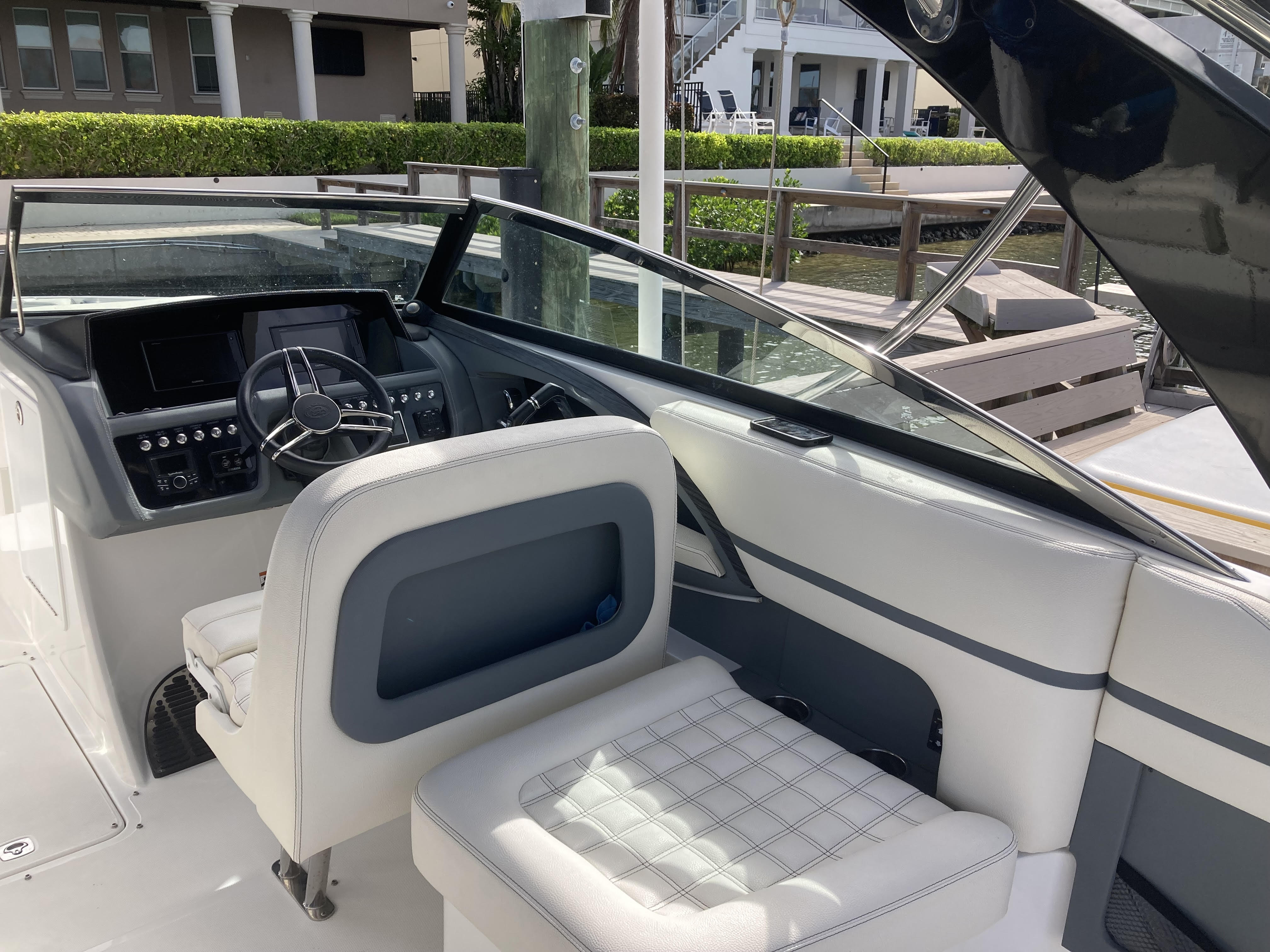 Sunlit white powerboat cockpit at a marina dock, featuring the helm with steering wheel and displays, quilted white passenger seat, glass windshield and wooden dock in the background.