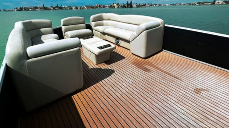 Sunny teak boat deck with white cushioned lounge seating on a pontoon, turquoise coastal waters and distant shoreline homes under a blue sky