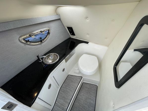 Compact marine head in a boat cabin with black countertop, stainless sink, white marine toilet, oval porthole, padded cream walls and mirrored door — small yacht bathroom.