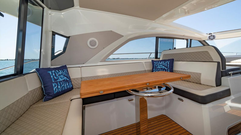 Cozy luxury yacht interior with U-shaped cream quilted seating, teak folding table, navy decorative pillows and panoramic windows framing calm blue ocean and distant shoreline.