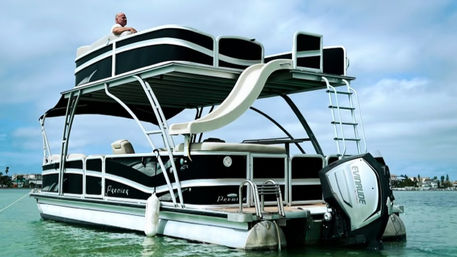 Double-decker pontoon boat with upper-deck water slide and outboard motor floating in turquoise coastal waters near a low shoreline under a cloudy sky, a person leaning on the top rail.