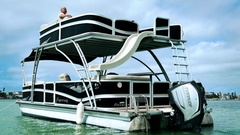 Double-decker pontoon boat with upper-deck water slide and outboard motor floating in turquoise coastal waters near a low shoreline under a cloudy sky, a person leaning on the top rail.