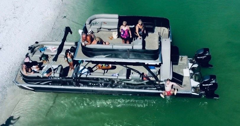 Aerial view of a black double-decker pontoon boat beached on a sandy sandbar in clear emerald-green water with a group of people relaxing and sunbathing.