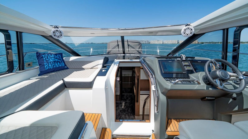 Sunlit luxury motor yacht helm and cockpit with cushioned lounge, blue decorative pillow, wooden steps down to the cabin, touchscreen navigation displays and panoramic ocean views under a clear blue sky.