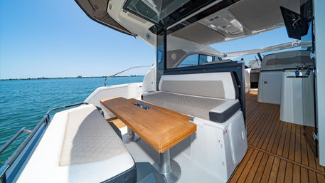 Sunlit luxury yacht seating area with teak table and quilted white-gray cushions on an open deck overlooking calm turquoise sea and clear blue sky