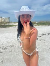 Person in a white bikini and rhinestone "BRIDE" cowboy hat smiling and showing an engagement ring on a sunny white-sand beach with blue sky and beachfront buildings in the background.