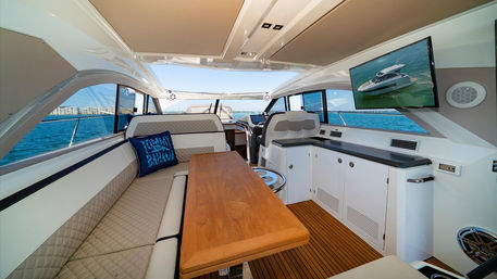 Sunny luxury yacht salon with teak dining table, cream quilted L-seat, built-in galley and wall TV, panoramic windows overlooking turquoise ocean and marina