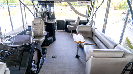 Spacious covered pontoon boat interior at a dock with tan leather wraparound seating, twin captain chairs, small teak table with cup holders, wet bar and lake views
