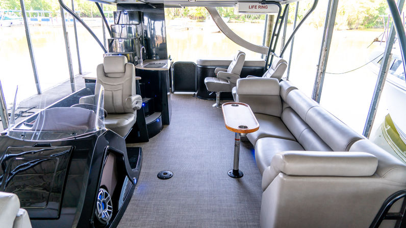 Spacious covered pontoon boat interior at a dock with tan leather wraparound seating, twin captain chairs, small teak table with cup holders, wet bar and lake views