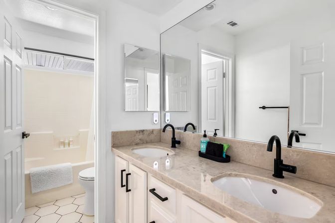 Bright modern white bathroom with double sink vanity, beige marble countertop, matte black faucets and large mirror, white cabinets, doorway to tub-shower with towel and toiletries, and hexagon tile floor.