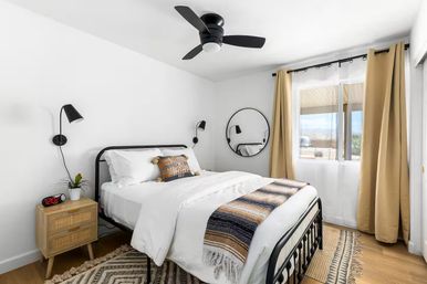 Bright modern bedroom with black metal bedframe, white bedding, striped woven throw, round wall mirror, wood nightstand, black wall lamps, tan curtains and ceiling fan